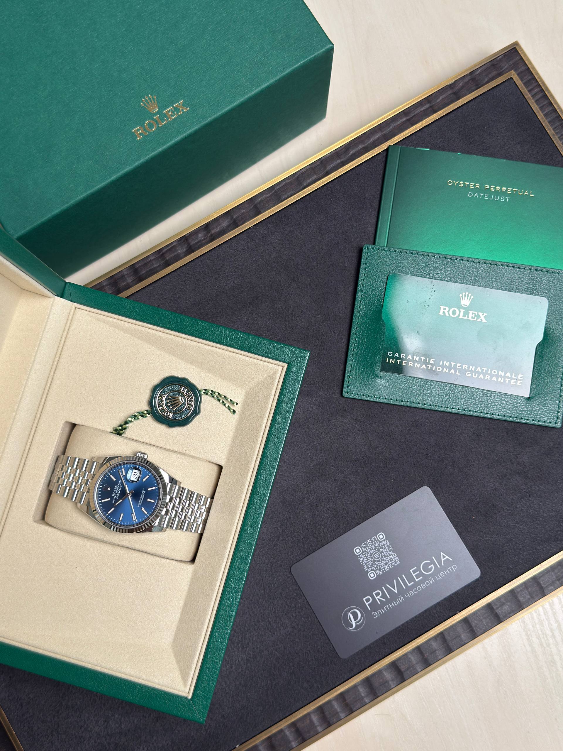 Green Rolex presentation box opened to reveal a silver bracelet watch with a blue dial and accompanying paperwork.