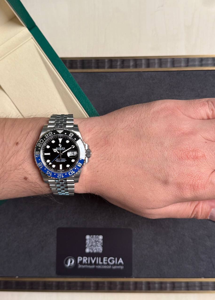 Wrist wearing a stainless steel Rolex GMT-Master II 'Batman' watch with blue and black bezel, resting on a dark presentation box with a Privilegia card nearby, indoors