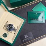 Green Rolex watch box opened to reveal a silver Rolex watch with black dial and blue‑red bezel in a cream interior, plus a green seal and cards nearby