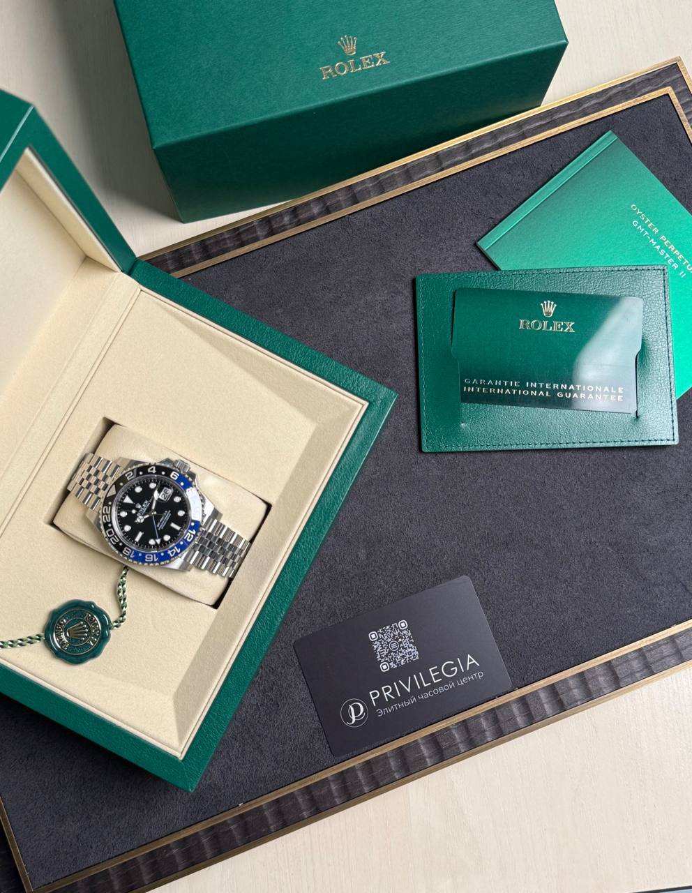 Green Rolex watch box opened to reveal a silver Rolex watch with black dial and blue‑red bezel in a cream interior, plus a green seal and cards nearby