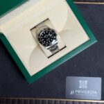 Rolex watch with black dial in a beige interior inside a green presentation box, metal bracelet visible.