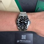 Silver Rolex Submariner wristwatch with black dial and magnified date window on a metal bracelet, worn on the left wrist; dark packaging box with 'PRIVILEGIA' in the background.