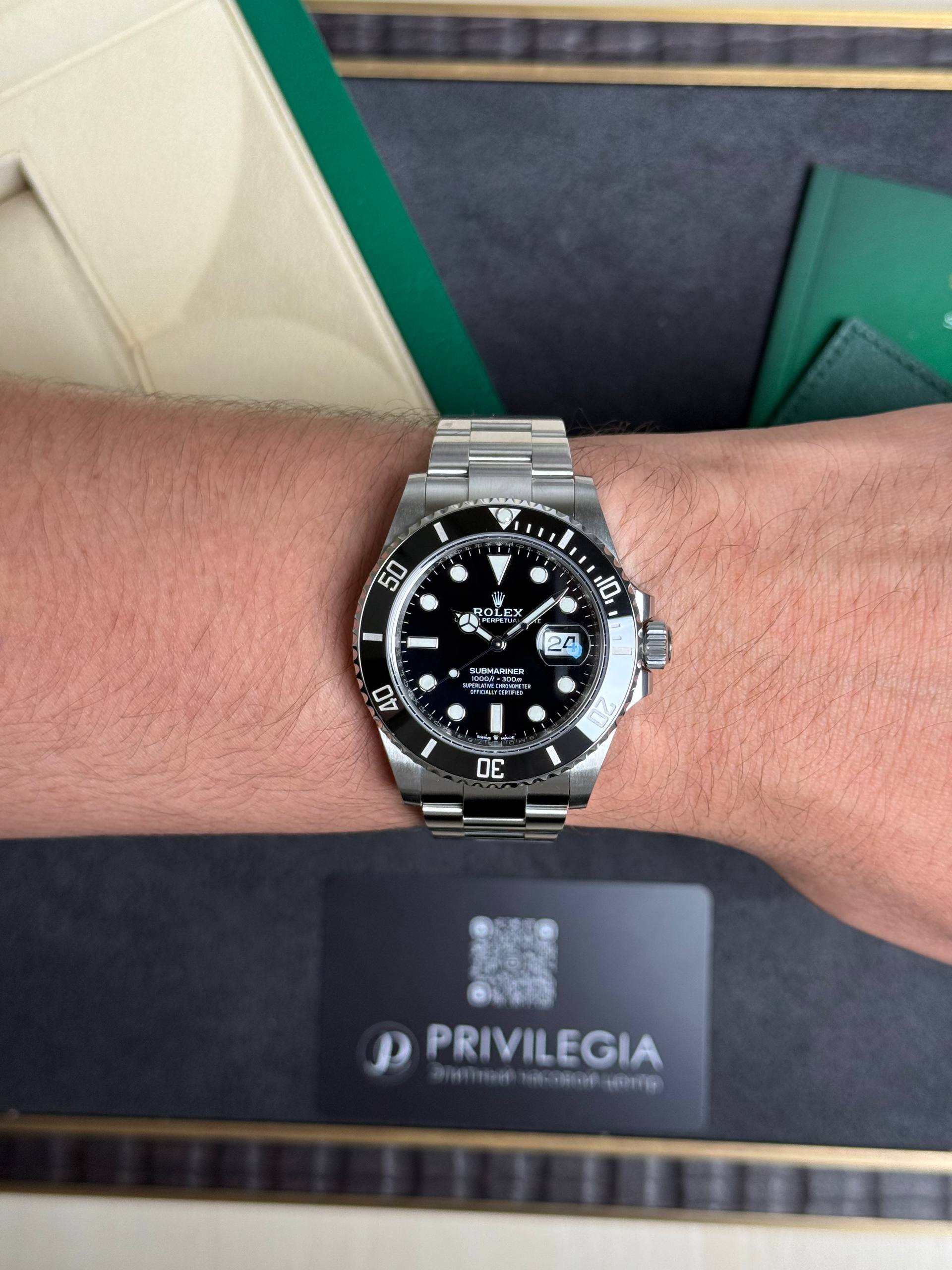Silver Rolex Submariner wristwatch with black dial and magnified date window on a metal bracelet, worn on the left wrist; dark packaging box with 'PRIVILEGIA' in the background.