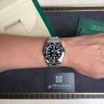 Wrist wearing a stainless steel Rolex Submariner diver watch, with green box and documentation in the background.