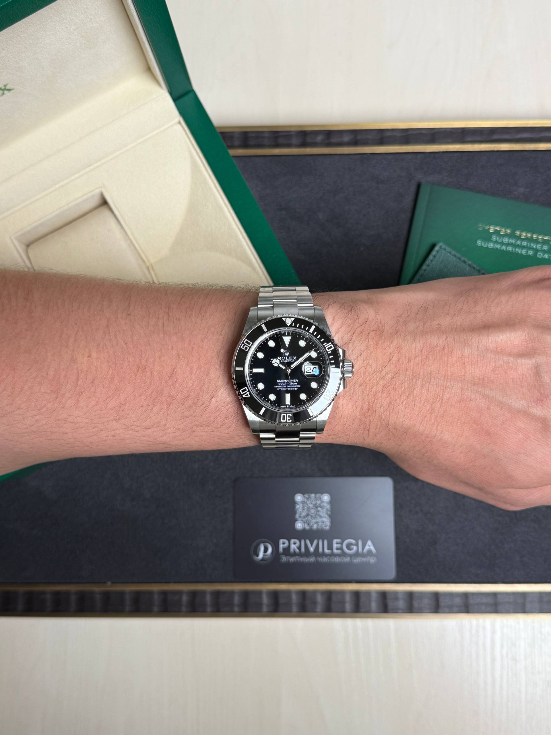 Wrist wearing a stainless steel Rolex Submariner diver watch, with green box and documentation in the background.