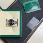 Rolex watch in a beige padded box with a green outer case, alongside a green warranty card and certification cards on a dark tray.