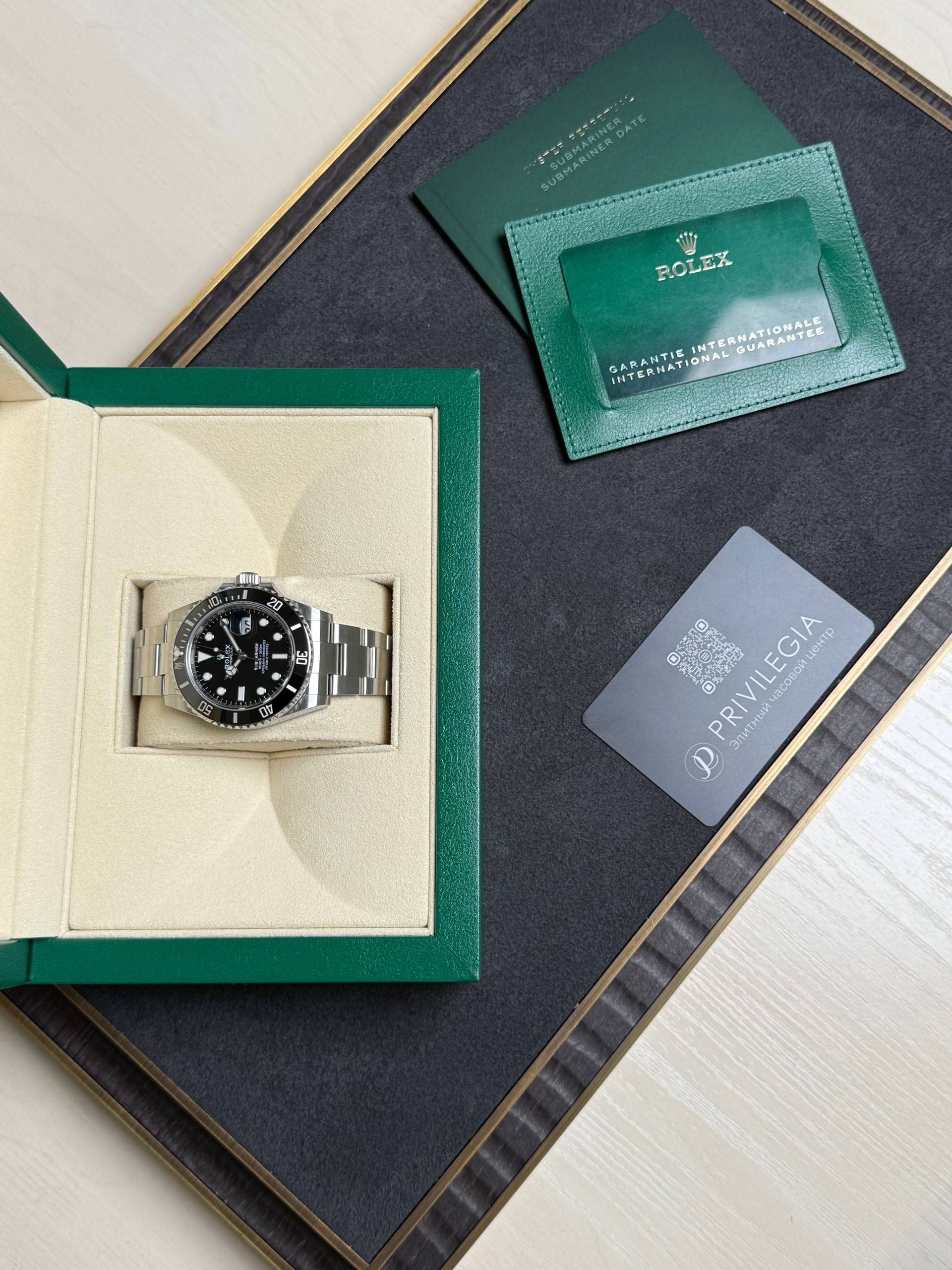 Rolex watch in a beige padded box with a green outer case, alongside a green warranty card and certification cards on a dark tray.