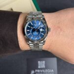 Rolex watch with blue dial on wrist, metallic band, close-up portrait-style shot with a black box in background