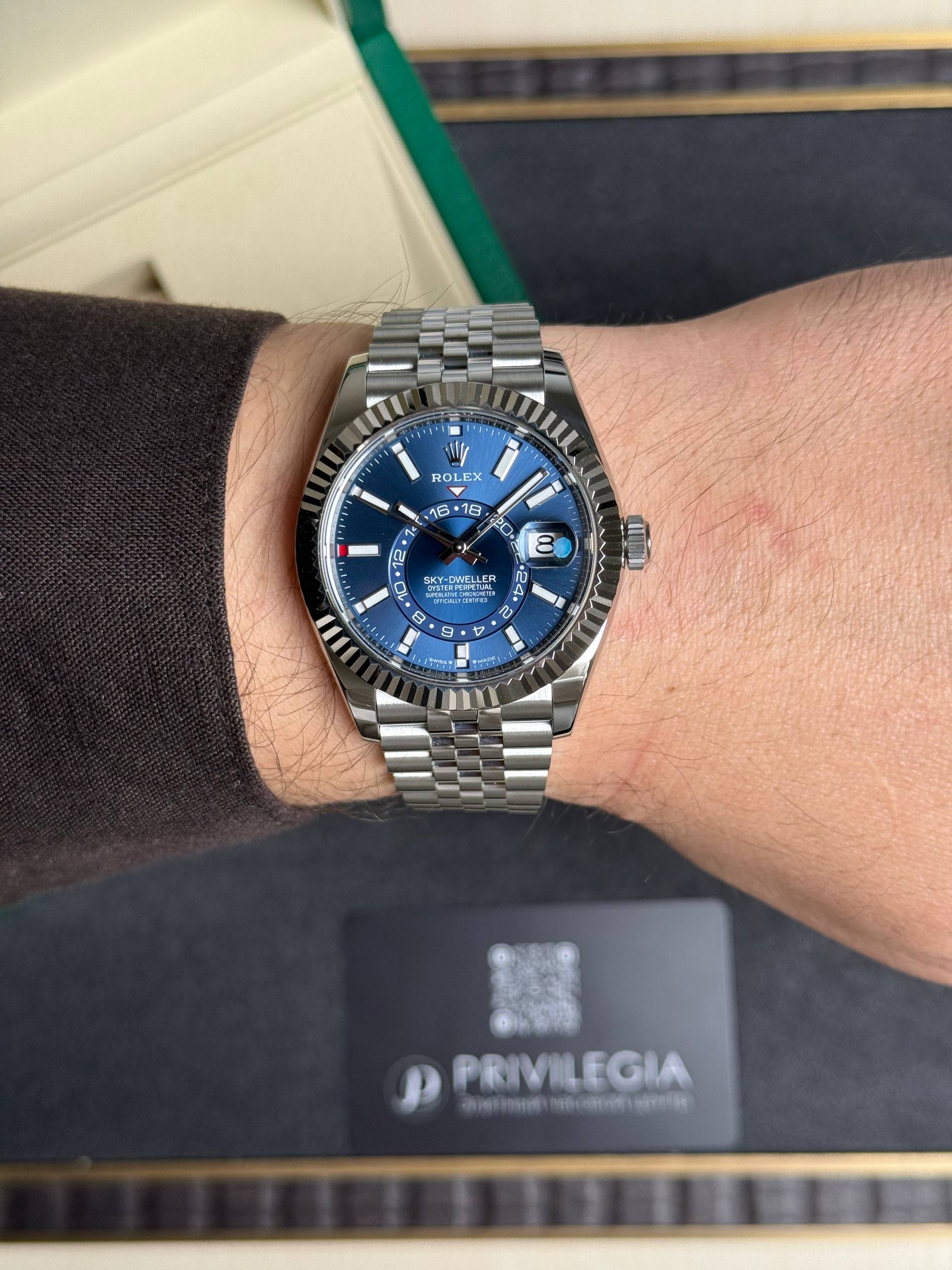 Rolex watch with blue dial on wrist, metallic band, close-up portrait-style shot with a black box in background