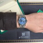 Wrist wearing a silver Rolex with a blue dial, next to an open green box and a Privilegia card on a dark surface.