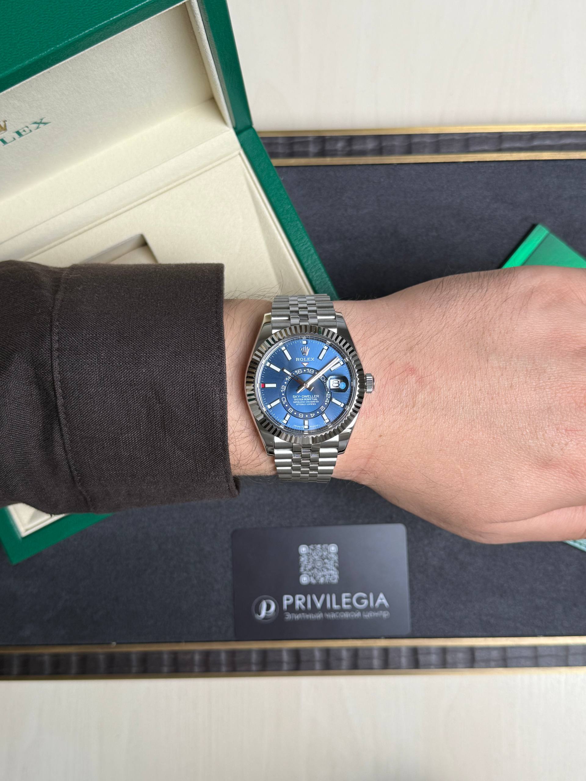 Wrist wearing a silver Rolex with a blue dial, next to an open green box and a Privilegia card on a dark surface.