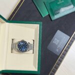 Rolex wristwatch in beige display box with green outer case and accompanying cards on a dark surface.