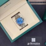 Rolex watch with a blue dial in an open cream interior/design box, silver bracelet visible.