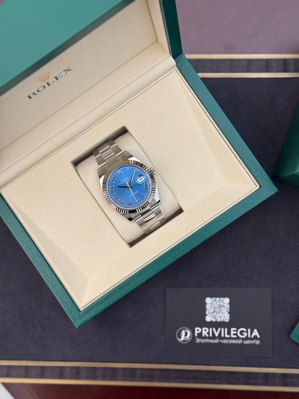 Rolex watch with a blue dial in an open cream interior/design box, silver bracelet visible.
