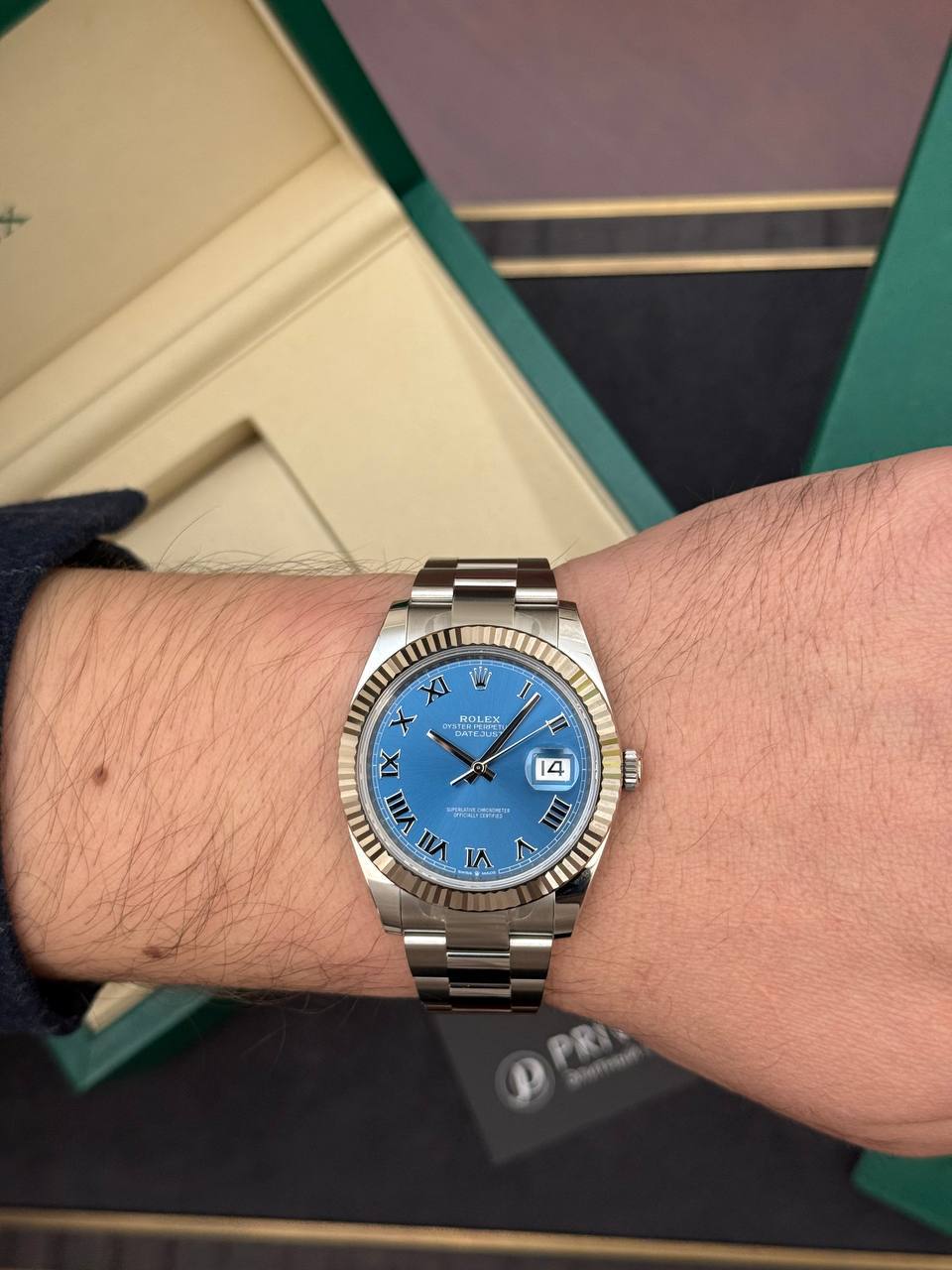 Wrist wearing a stainless steel Rolex Datejust with a blue dial, Roman numeral hour markers, and a date window at 3 o'clock.