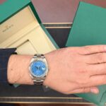 Wrist wearing a silver Rolex watch with a blue dial and Roman numerals, open green box beside it.