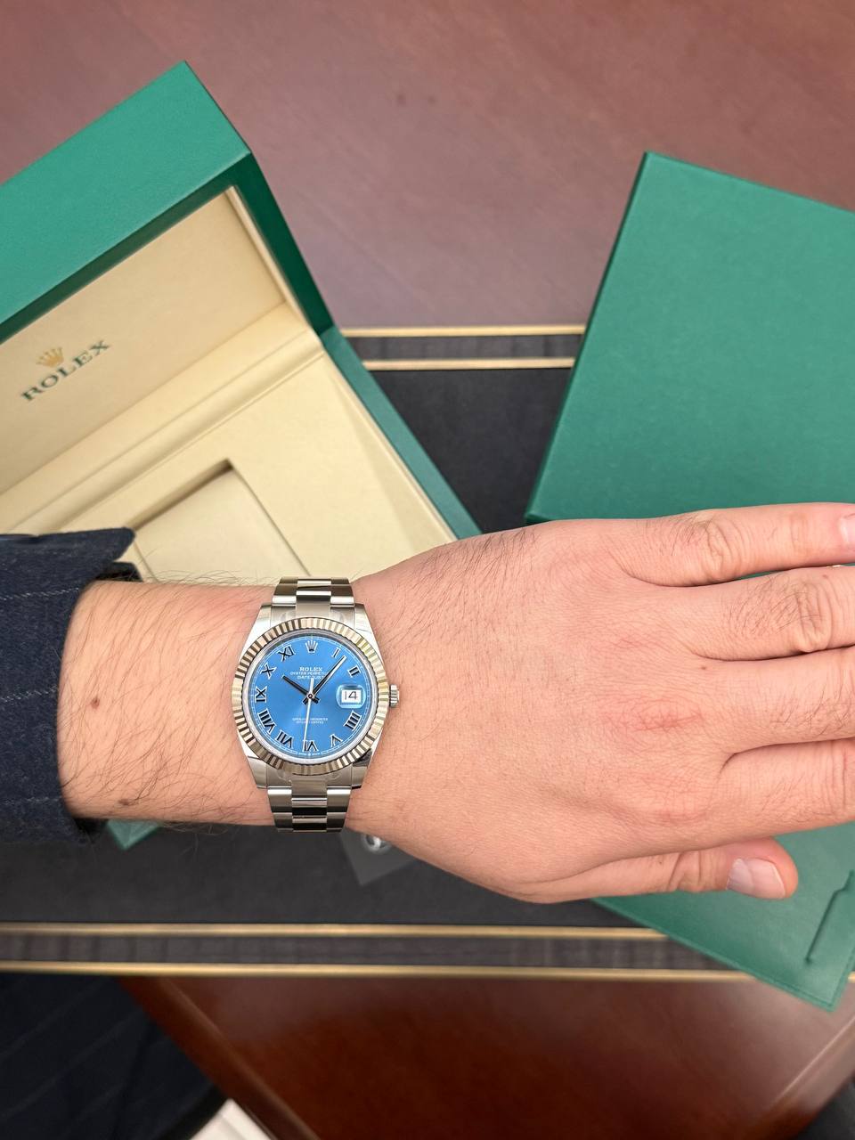 Wrist wearing a silver Rolex watch with a blue dial and Roman numerals, open green box beside it.