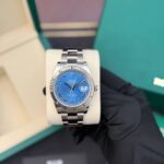 Rolex Datejust stainless steel watch with blue dial and Roman numerals on a display cushion, held by a gloved hand.
