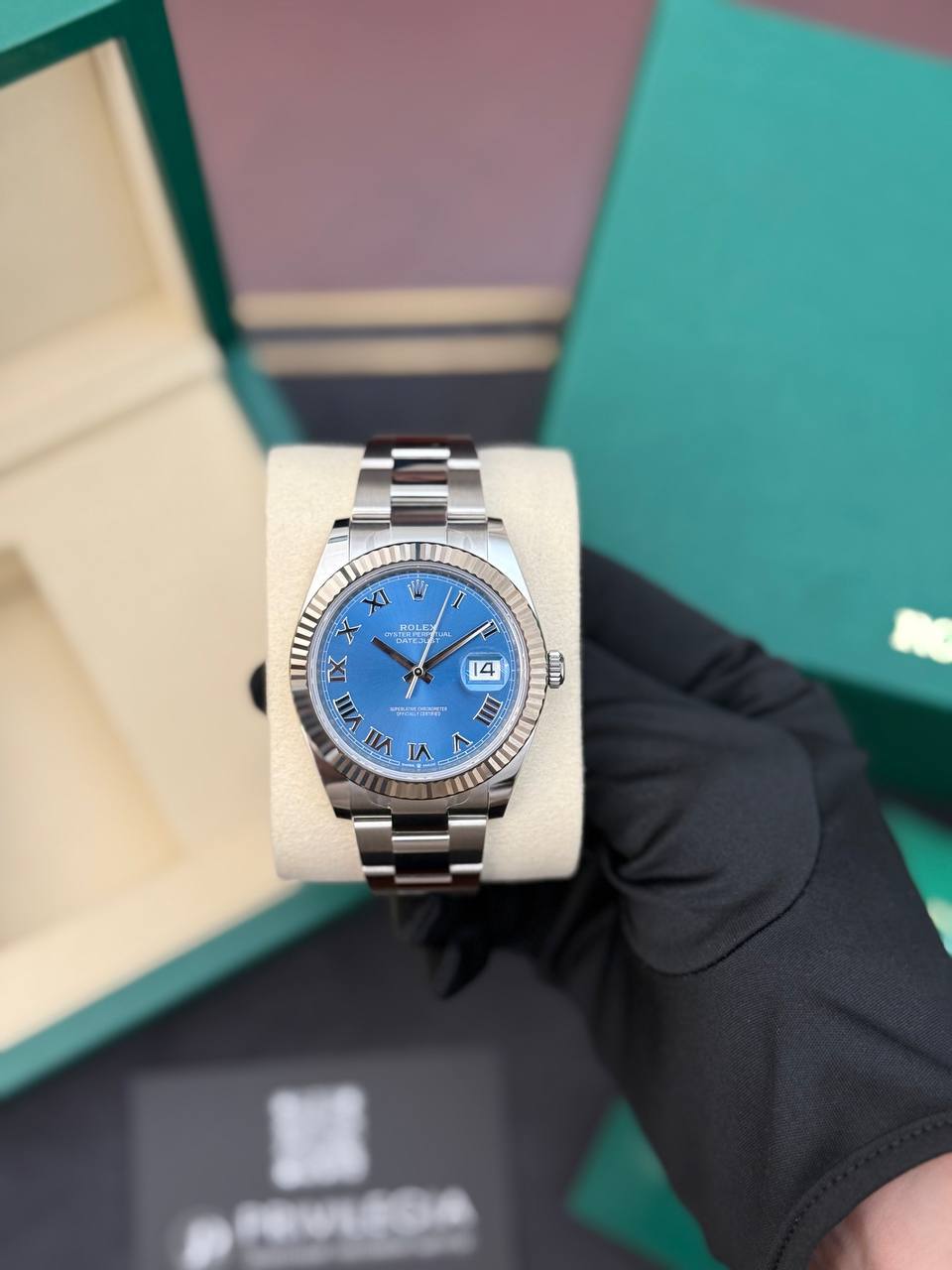 Rolex Datejust stainless steel watch with blue dial and Roman numerals on a display cushion, held by a gloved hand.