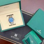 Rolex watch with a blue dial displayed inside an open green presentation box, surrounded by green warranty cards and booklets.