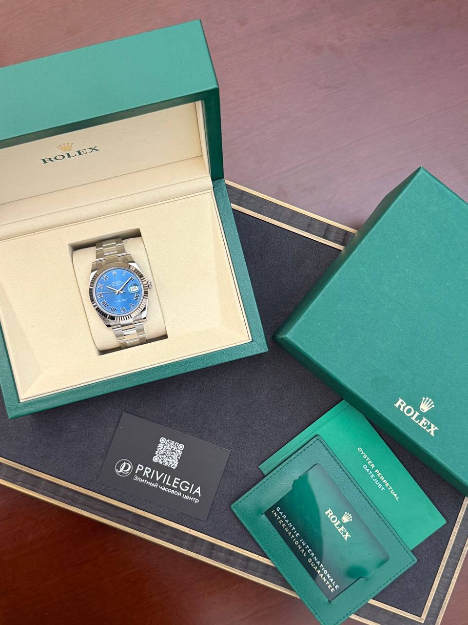 Rolex watch with a blue dial displayed inside an open green presentation box, surrounded by green warranty cards and booklets.