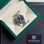 Open green Rolex box reveals a stainless steel Datejust on a beige cushion with Roman numerals and fluted bezel.