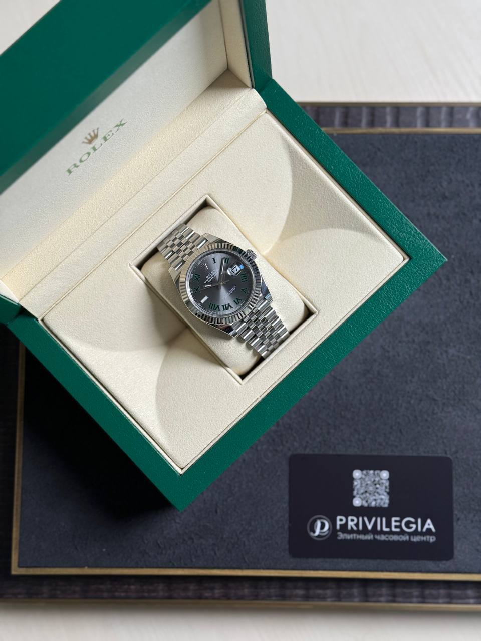 Open green Rolex box reveals a stainless steel Datejust on a beige cushion with Roman numerals and fluted bezel.