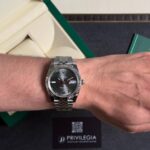 Silver Rolex Datejust on the wrist with a fluted bezel and green Roman numeral markers; date window at 3 o'clock. Box and Privilegia card visible in the background.