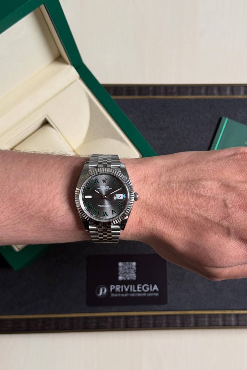 Silver Rolex Datejust on the wrist with a fluted bezel and green Roman numeral markers; date window at 3 o'clock. Box and Privilegia card visible in the background.