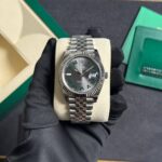 Rolex watch with a silver Jubilee bracelet resting on a cream display pillow, green box in the background.