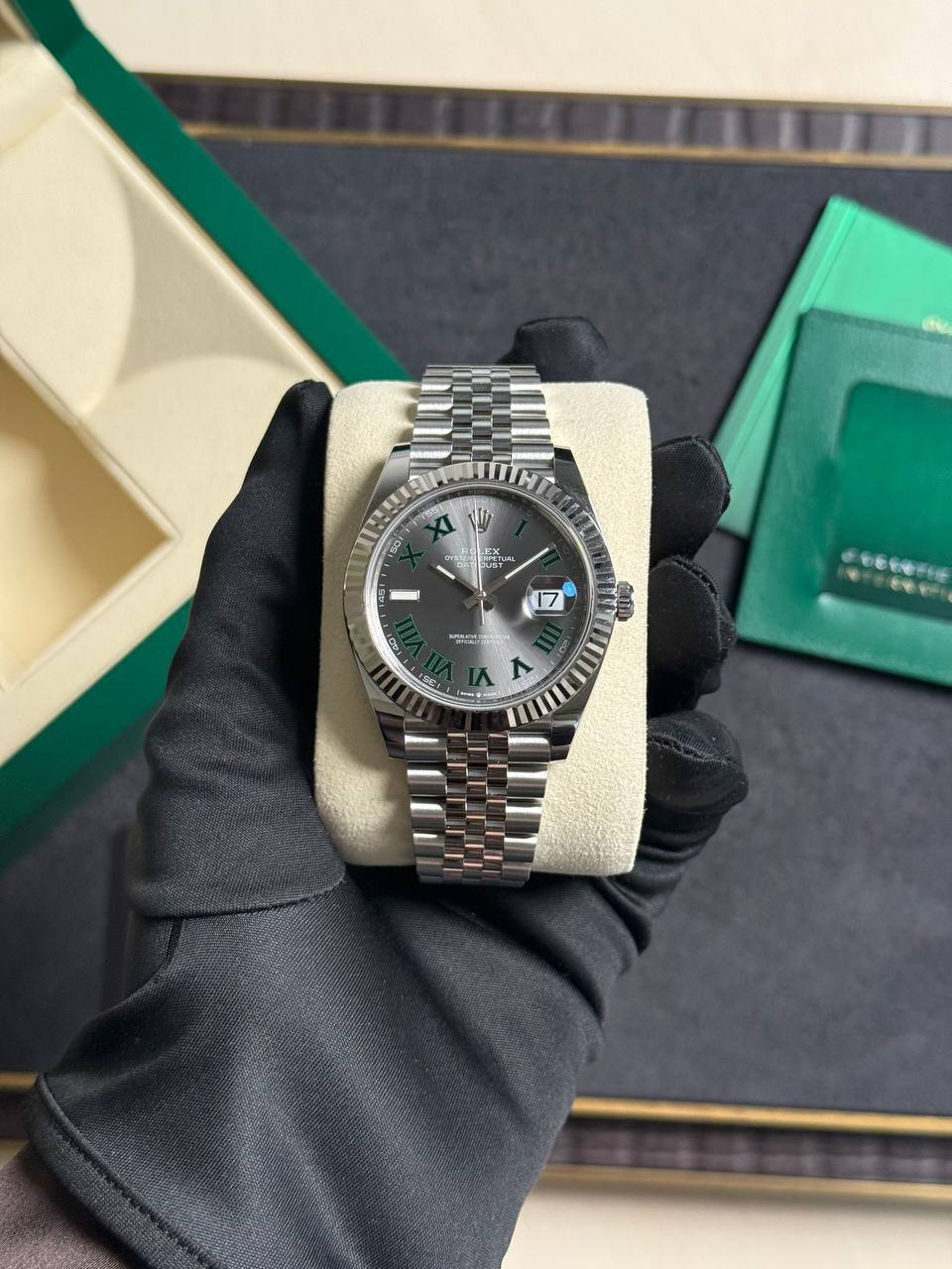 Rolex watch with a silver Jubilee bracelet resting on a cream display pillow, green box in the background.