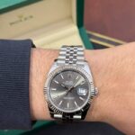 Stainless steel Rolex Datejust on the wrist with a green Rolex box in the blurred background, showing the date window at 3 o'clock.