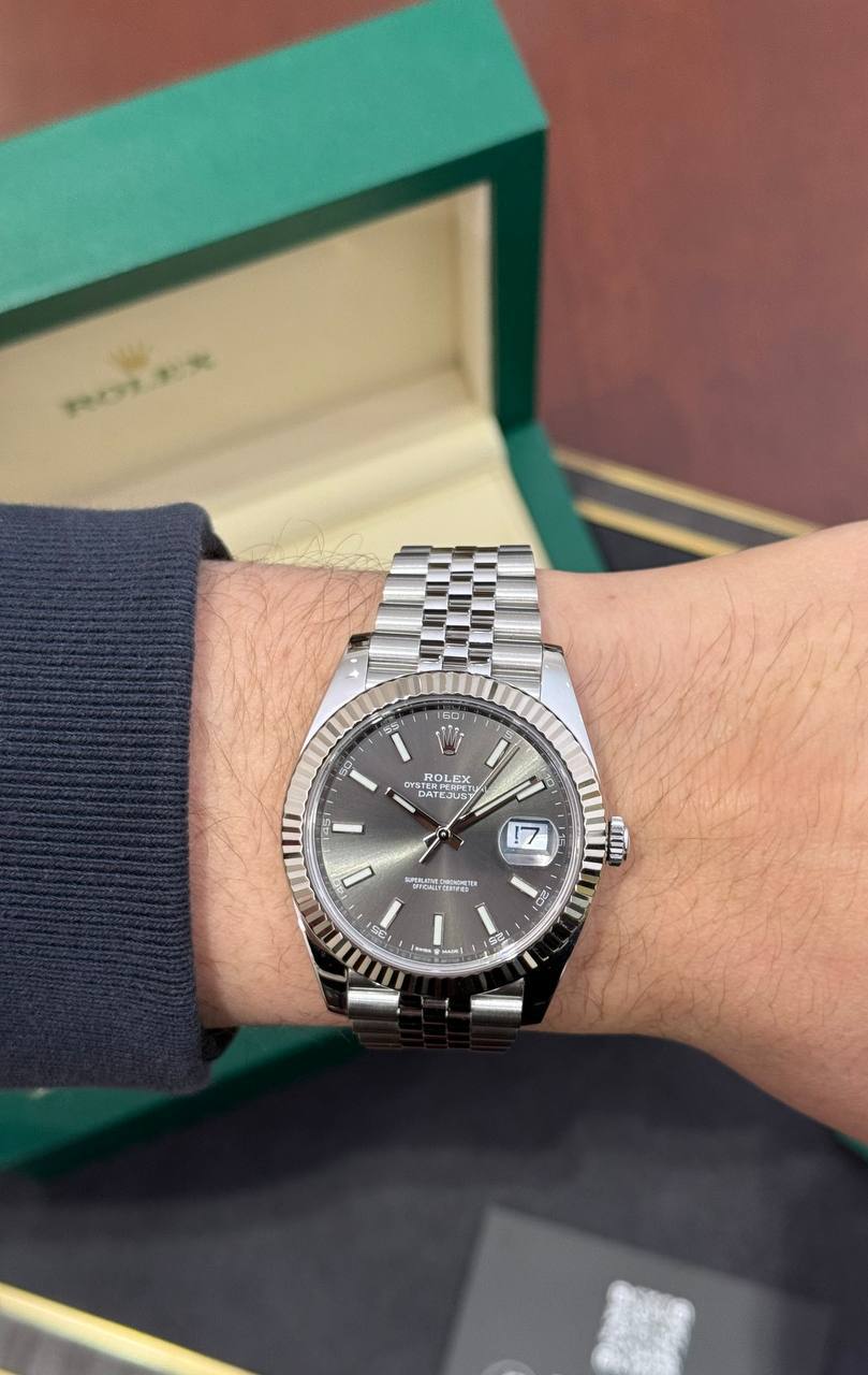 Stainless steel Rolex Datejust on the wrist with a green Rolex box in the blurred background, showing the date window at 3 o'clock.