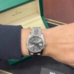Wrist wearing a stainless steel Rolex Datejust with silver dial and date window, next to an open green Rolex box.