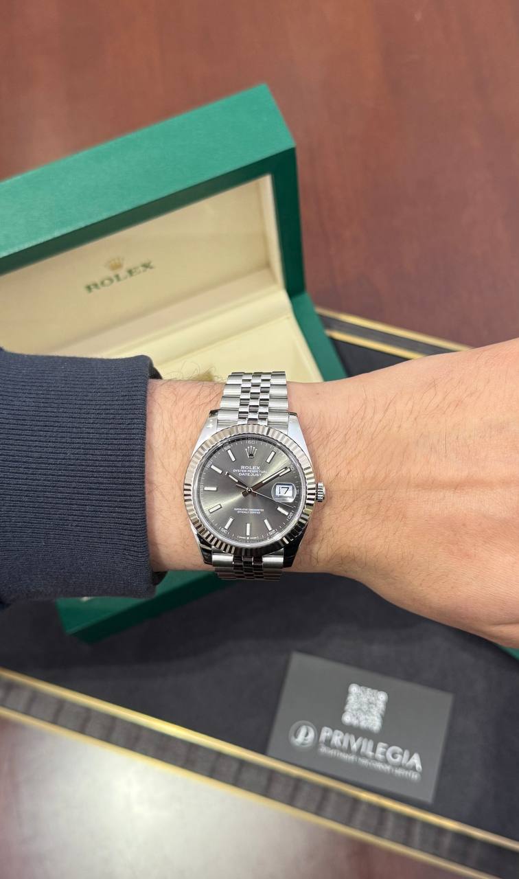 Wrist wearing a stainless steel Rolex Datejust with silver dial and date window, next to an open green Rolex box.