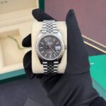 Silver Rolex Datejust watch on a beige cushion, held by a gloved hand, with green box in the background.