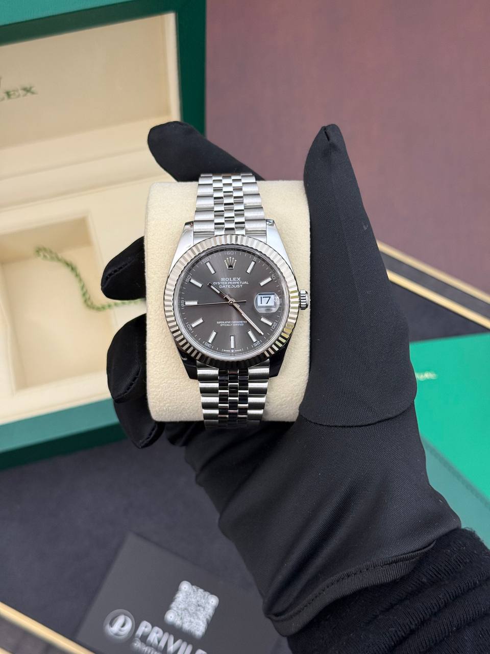 Silver Rolex Datejust watch on a beige cushion, held by a gloved hand, with green box in the background.