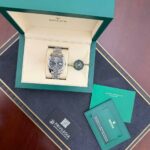 Rolex watch in open green box with beige interior and accompanying green card and booklet on a dark surface (product photography)