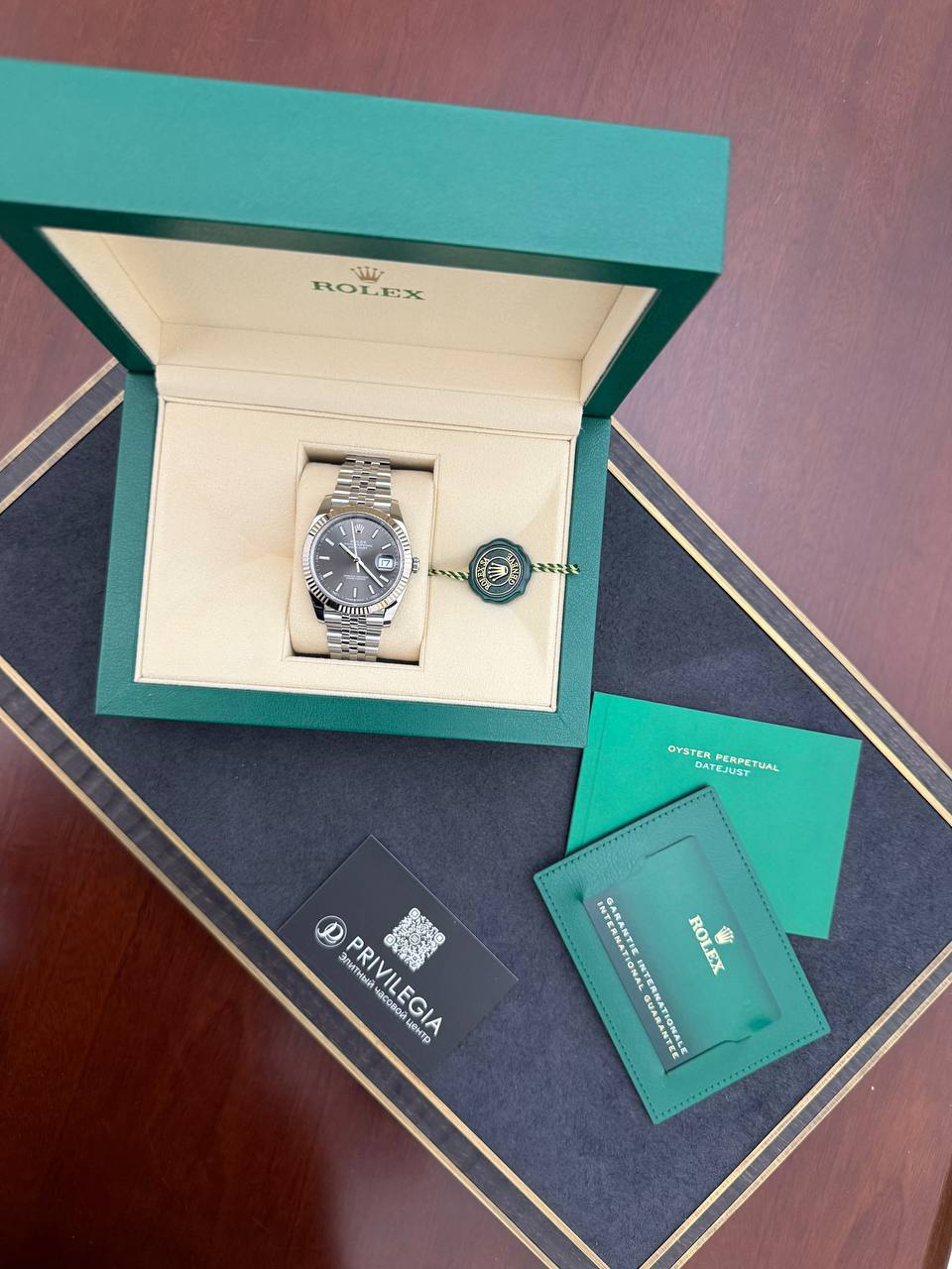 Rolex watch in open green box with beige interior and accompanying green card and booklet on a dark surface (product photography)