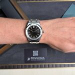 Stainless steel wristwatch on the forearm with a black textured dial; packaging and a Privilegia card visible in the background.