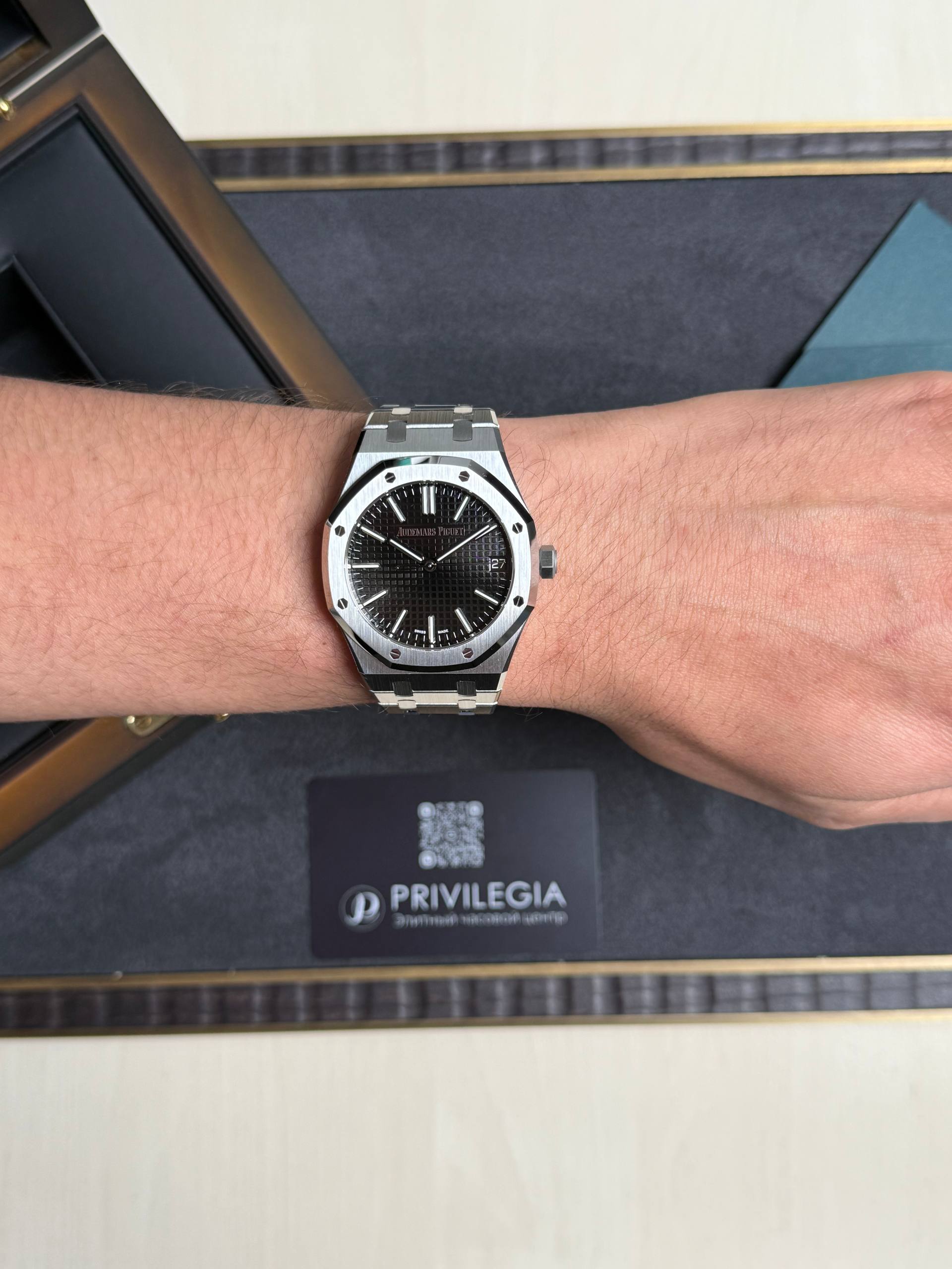 Stainless steel wristwatch on the forearm with a black textured dial; packaging and a Privilegia card visible in the background.