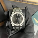 A stainless steel Audemars Piguet Royal Oak watch on a cushioned display held by gloved hands, showing a textured dark dial and date window at 3 o'clock.