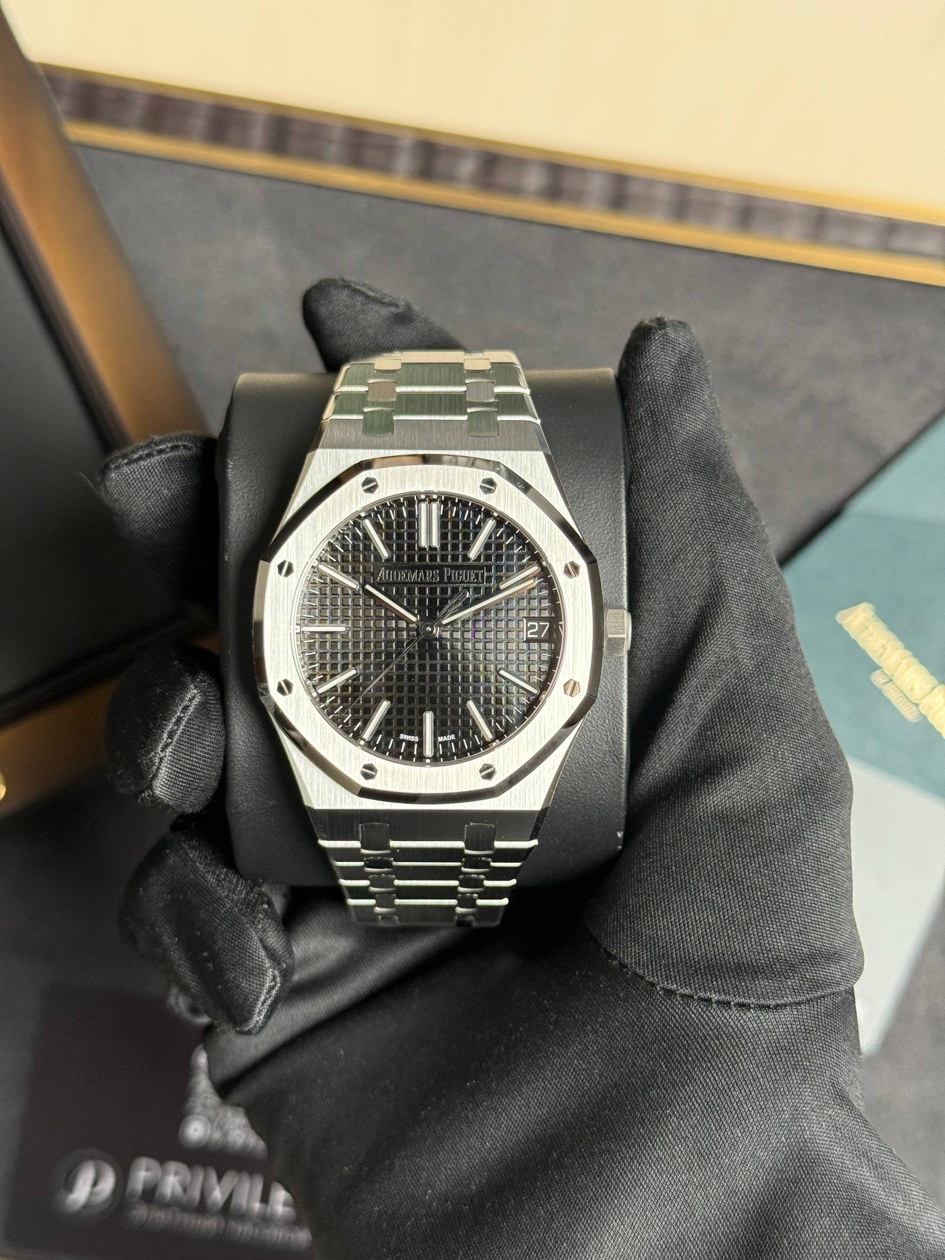 A stainless steel Audemars Piguet Royal Oak watch on a cushioned display held by gloved hands, showing a textured dark dial and date window at 3 o'clock.