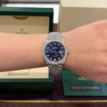 Rolex Datejust on a wrist: blue dial with diamond bezel and date window, boxed in green Rolex packaging in the background.