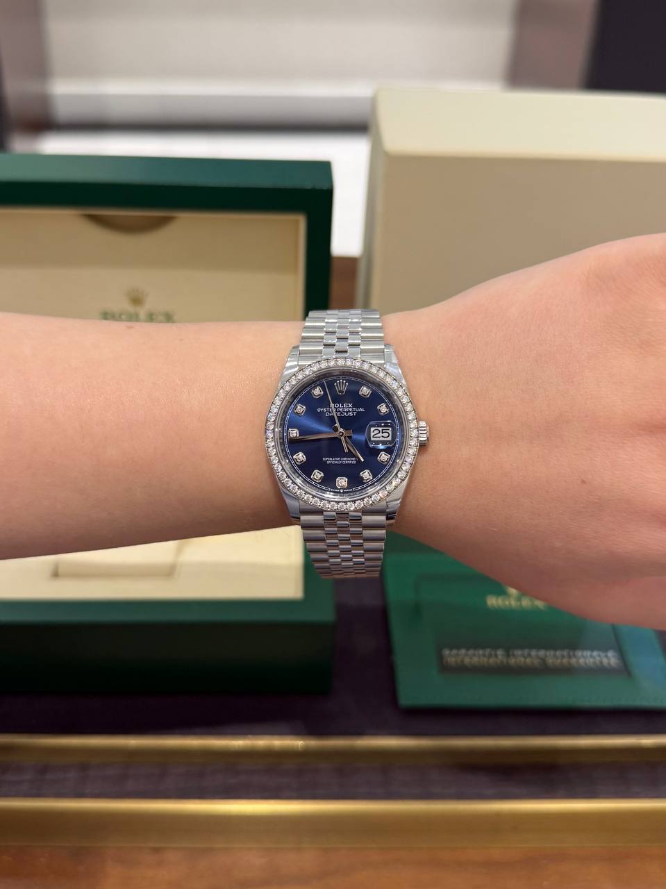 Rolex Datejust on a wrist: blue dial with diamond bezel and date window, boxed in green Rolex packaging in the background.