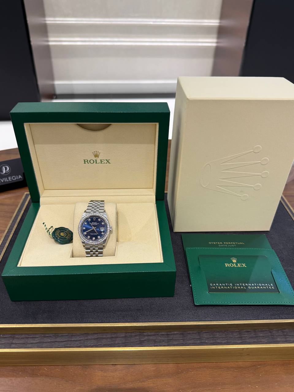 Rolex Oyster Perpetual Datejust in green presentation box with blue dial and diamond bezel, displayed on a store counter.