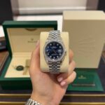 Hand holding a silver Rolex Datejust with a blue dial and diamond bezel, in front of its green box and beige display cushion.