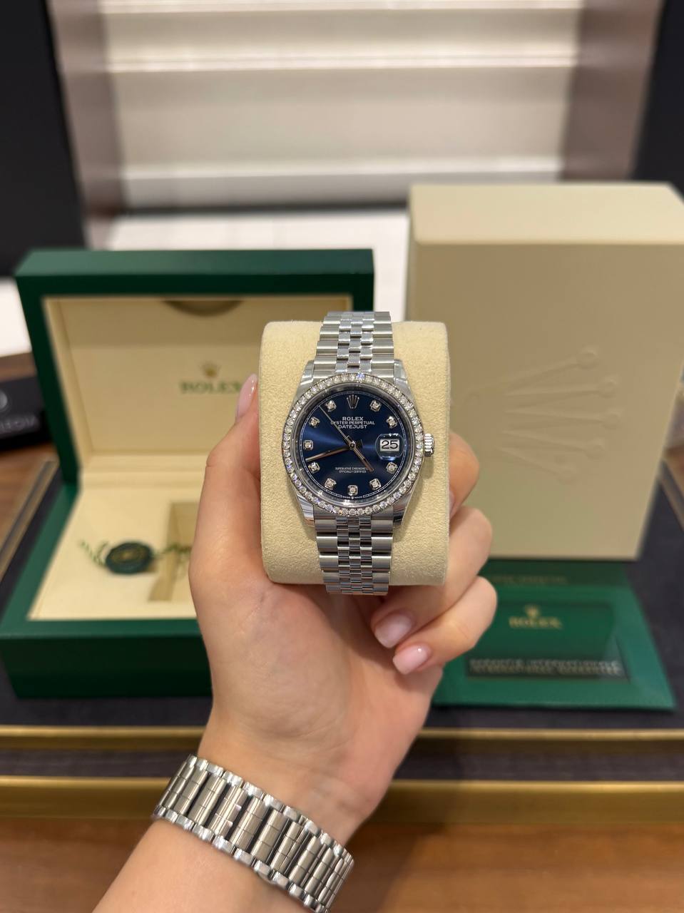 Hand holding a silver Rolex Datejust with a blue dial and diamond bezel, in front of its green box and beige display cushion.