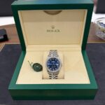 Rolex watch with blue dial and diamond bezel in an open beige interior of a green presentation box with a green tag beside it.