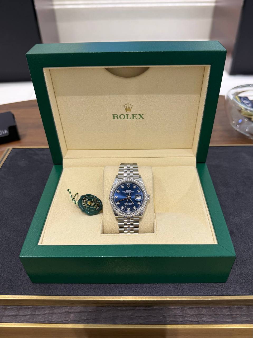 Rolex watch with blue dial and diamond bezel in an open beige interior of a green presentation box with a green tag beside it.